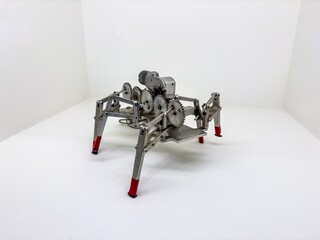 This image features a close-up view of a mechanical spider robot prototype with a visible gear mechanism and metallic structure. The robot has four legs with red-tipped feet, designed for balance and