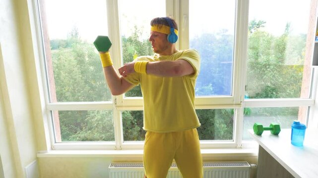 Comic athlete with dumbbells pumps a muscle while listening to music on headphones. A young funny guy training at home. Workout with dumbbells. Slow motion.
