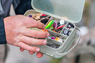 The fisherman's hands are holding a plastic box with a variety of lures and fishing hooks