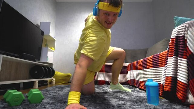 Comic funny athlete at home pumping his biceps while listening to music on headphones. A young funny guy training at home. Workout push-ups with hands. Slow motion.