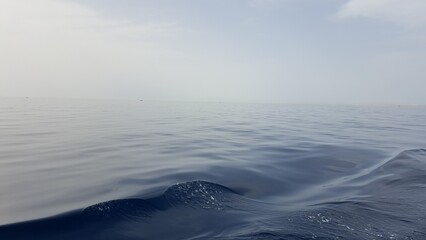 A serene, vast expanse of deep blue ocean water stretches out to the horizon under a soft, hazy sky, captured by a smooth, rippling wave close to the camera, calming and peaceful maritime scene