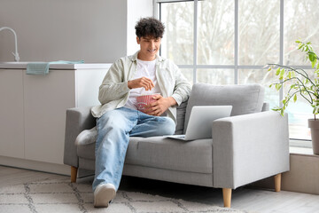 Young man with popcorn and laptop on sofa in kitchen