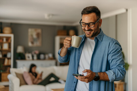 Young professional freelancing at home, texting on a phone in a stylish, well-lit living room. A woman enjoying on the sofa in the background