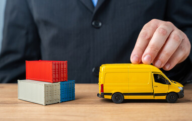 A businessman pushes a delivery van past a stack of shipping containers. The concept of delivery...