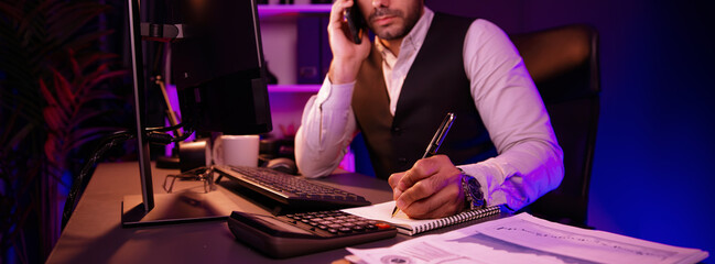 Smart businessman calling partner or investor on smartphone on panoramic view, writing data on paper work report to analyze sales channel discipline's product service at neon dark light room. Surmise.