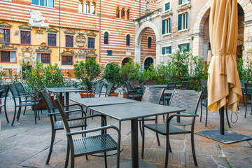 Empty outdoor cafe tables and chairs outside in old town square Piazza dei Signori, Verona city, Veneto, Italy. Italian square with sidewalk restaurant and colorful buildings. Tourist destination