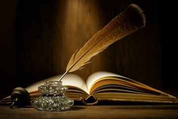Open antique book with feather and ink bottle on old wooden table. Feather for calligraphy, old...