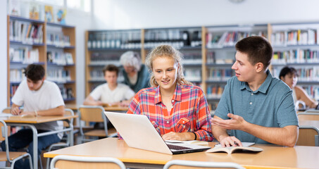 Teenage students help each other study at laptop while sitting in the school library
