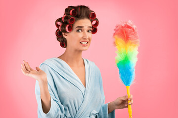 Spring Cleaning Concept. Confused Housewife With Curlers Holding Feather Duster Standing Over Yellow Background. Studio Shot