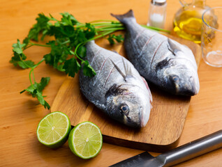 Two gilt-head breams laid on wooden cutting board with lime herbs salt and oil