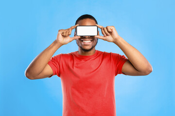 Black Man Holding Phone With Blank Screen In Front Of Eyes, Looking At You Through His Smartphone Over Blue Studio Background. Guy Posing With Cellphone Advertising New Mobile Application