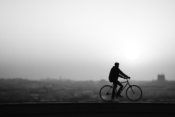 Obraz premium Silhouette of a cyclist riding a bicycle against a hazy city backdrop at sunset.