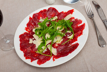 Thin slices of meat carpaccio, cheese and arugula on white plate served on table in restaurant. Traditional Italian dish