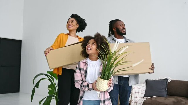 Happy joyful african american dad mom and their preschool cute daughter, moved to their new apartment, holding boxes with home things, girl holding a flower pot, they come in at home in excitement