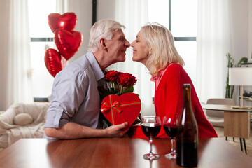elderly senior couple celebrating valentine's day at home and kissing, old man giving his wife gift, flowers and heart shaped balloons, retired people in love