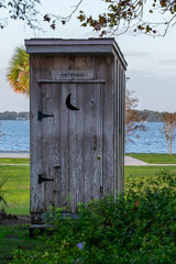 A vintage grey weathered and worn wooden outhouse with a carved out crescent moon. The privy latrine is on a green grassy meadow with water in the background. The old lavatory door is closed.