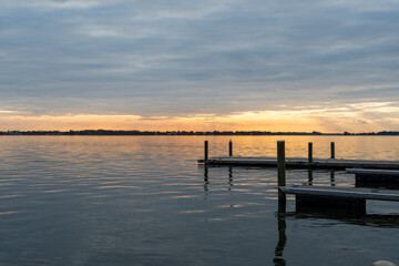 Fototapeta premium A dramatic sunset with orange and yellow hues at the horizon. The sky is filled with thick clouds reflecting on the calm water. There’s a floating boat dock with mooring posts. The jetty is wooden.