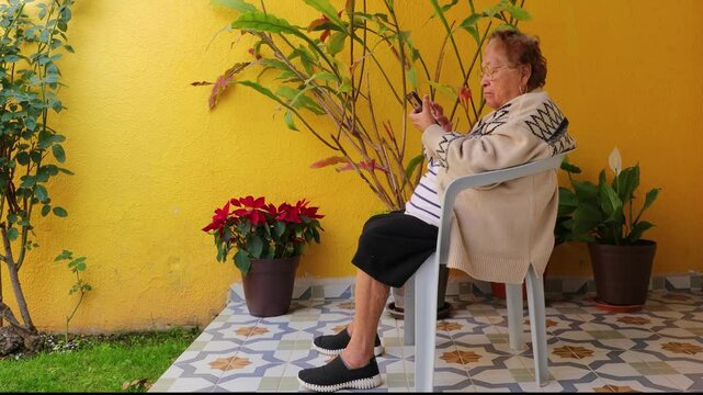 Senior woman watching and playing with mobile phone sitting on chair in garden, very concentrated with serious expression. Concept of communication and being connected in adulthood