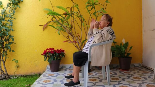Senior woman exercising arms by raising and lowering arms in coordinated manner, sitting on chair in garden, yellow wall in background. Concept of home exercise for seniors and healthy lifestyle