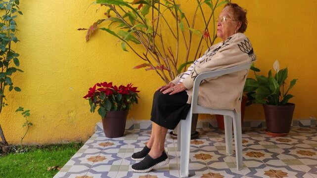 Senior woman exercising her legs sitting on a chair in garden, raising and lowering her heels, yellow wall in background. Concept of exercises for seniors at home and healthy lifestyle