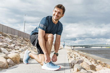 Man tying shoelaces while jogging on riverbank during partly cloudy afternoon