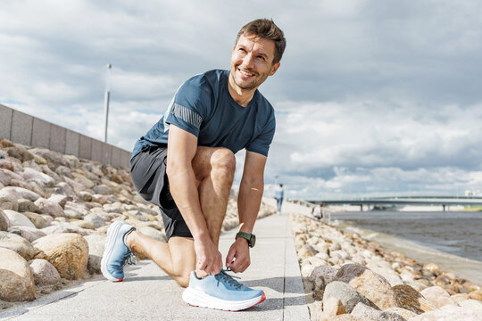 Man tying shoelaces while jogging on riverbank during partly cloudy afternoon