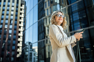 Obraz premium Businesswoman using smartphone while standing outside modern office building on sunny day