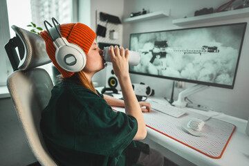 Young gamer girl wearing vibrant orange cap and white headphones sits in stylish gaming chair,...