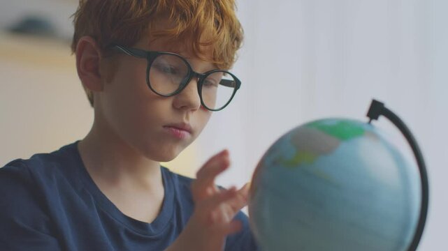 A boy with ginger hair and glasses is focused on a globe while engaging in remote learning. He is exploring different countries and honing his geography skills from his study area.