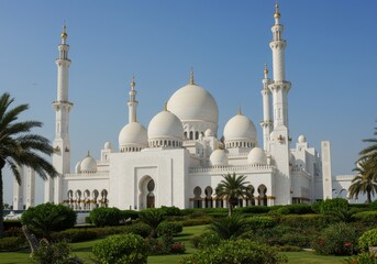 A majestic white mosque with turquoise domes and tall minarets, surrounded by lush green tropical plants and palm trees