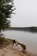 Mother and child sitting by the lake. Woman in white dress holding child sitting on grassy lake shore with forest in background. Peaceful nature concept