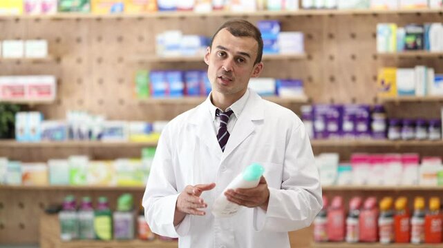 Polite young male apothecary holding shampoo standing against shelves full of medicine in drugstore 