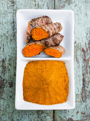 Turmeric rhizomes and powder resting in a white ceramic bowl on rustic wooden table