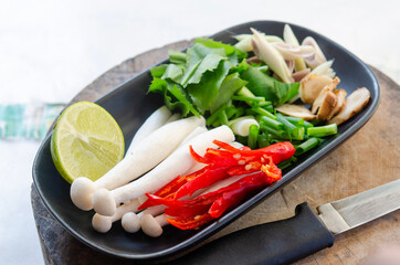 Fresh Ingredients for Tom Yum Soup on a Black Plate: Lime, Lemongrass, Galangal, Red Chili, Mushrooms, and Herbs, with a Knife on a Wooden Cutting Board