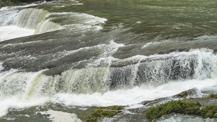 A fragment of a riverbed. Stormy streams foam on the ledges of the rapids. Green grass on the rocks. Splashes. China. Silver Chain Waterfall. Guizhou