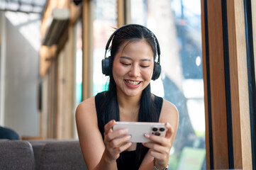 A cheerful Asian woman enjoys playing a mobile game while relaxing in a cafe.