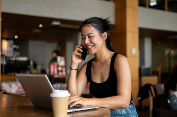 A confident Asian woman is talking on the phone while working on her laptop, working remotely.