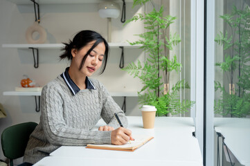 A beautiful, calm Asian woman writes in her diary while relaxing in a minimalist cafe.