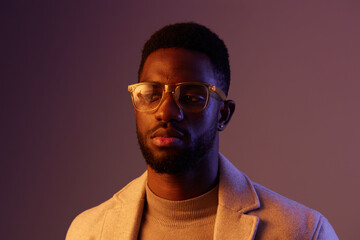Portrait of a thoughtful Black man wearing stylish glasses and a beige coat, with soft lighting creating a warm and modern atmosphere