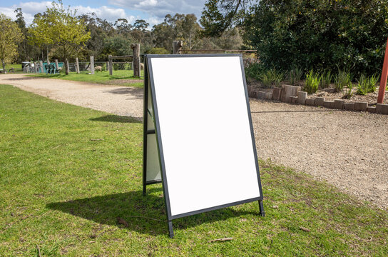 a blank A-frame sandwich board placed on a grassy area in an outdoor park.Empty white background texture with copy space to add custom text or graphics,for promoting events, directions, or information