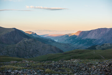 Fototapeta premium Scenic top view from green grassy stony pass to golden mountains, illuminated by setting sun under cloud in clear sunset color sky. Mountain range far away in soft sunset tones. Gold light on hills.