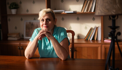 A smiling mature woman with short blonde hair sits at a wooden table, resting her hands under her chin. The background features bookshelves, creating a cozy and peaceful indoor atmosphere.