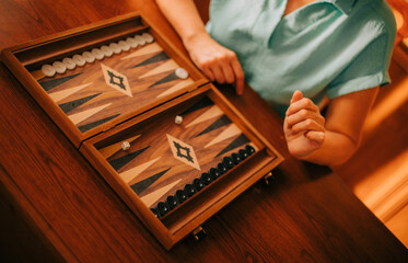 A close-up of a woman playing backgammon at home, with dice and game pieces on the wooden game...