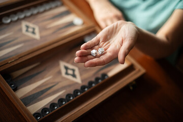 A woman is playing backgammon, holding two dice in her hand while focused on the game. The wooden backgammon board is laid out on the table with black and white pieces, reflecting casual indoor play.