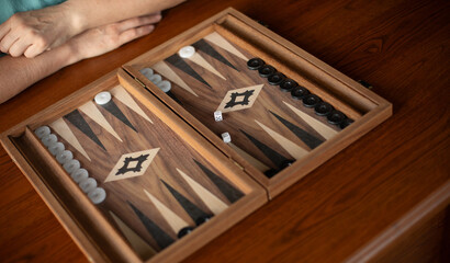 A close-up of a wooden backgammon board with dice and game pieces, showing a person’s hand...