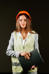 Woman engineer holding laptop while wearing safety gear against a black background