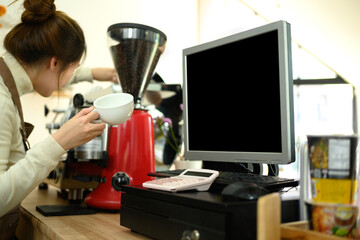 Computer monitor and cash register on wooden counter with barista preparing a drink in background