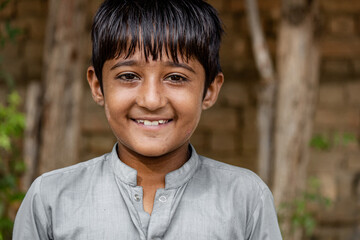 Smiling young boy with short black hair, wearing a traditional gray shirt, standing outdoors against a rustic background with trees and a brick wall.

