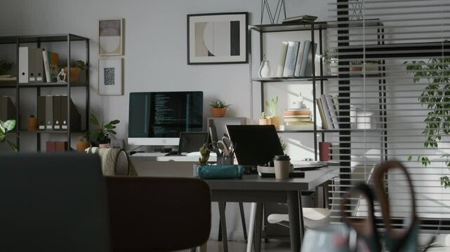 No people view of modern office of IT company with computers on desks, two racks with file folders and books on shelves, abstract paintings on wall
