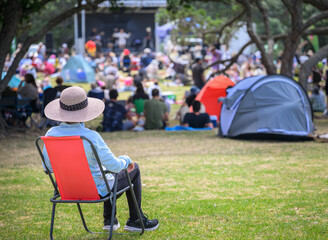 Woman sitting on camping chair, unrecognizable crowds listen to the music concert in the park....